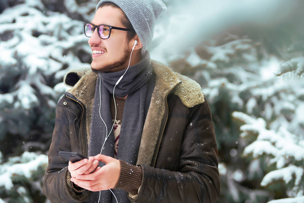 Young man with phone in winter park.  Businessman uses a phone and  enjoying winter moments in a snowy forest. Snowfall. - Foto, imagen