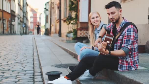 Mladá dvojice pouličních zpěváků sedících na chodníku, hrajících na kytaru a zpívajících - Záběry, video