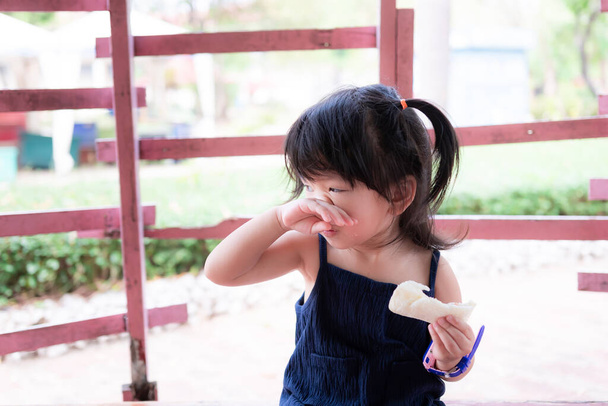 Söpö tyttö murskattu nenä oikealla ja vasemmalla kädellä pitää valkoinen voileipä. Pieni lapsi kutiava nenä pöly allergia oireita. Lapset hierovat nenäänsä takakäsiin. Suloinen lapsi on 2-vuotias. - Valokuva, kuva