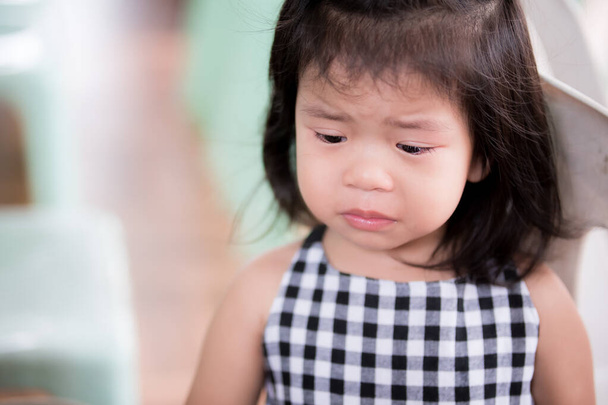 Little girl made a sad face from regret. Asian children bend their heads to cry. There are tears in eyelashes. Child frowns from uneasiness. Kid is wearing black and white checkered shirt. 3 years old - Photo, Image