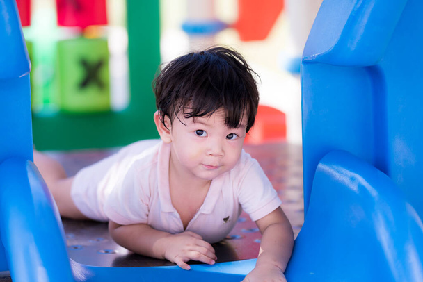 Przystojny mały azjatycki chłopiec bawić się na placu zabaw w upale lata lub wiosny. Dziecko patrzy w kamerę i uśmiecha się lekko. Maluch poci się na twarzy. Dziecko ma 2 lata.. - Zdjęcie, obraz