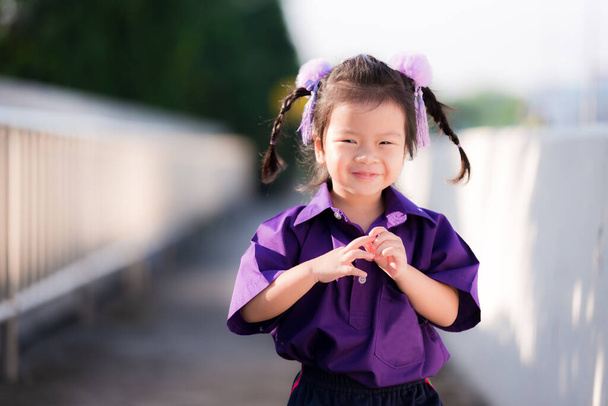 Cute child shows blushing expression when being photographed. Sweet smiley girl wearing purple dress. The sun shines in the evening. Asian kid aged 3 year old. Preschooler wear colored sports uniform. - Photo, Image