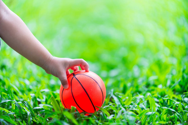 Kleine Mädchenhand wird für einen orangefarbenen Ball vom grünen Rasen gehalten. Kind spielt Gummiball. Sinneskonzepte und Lernen. - Foto, Bild