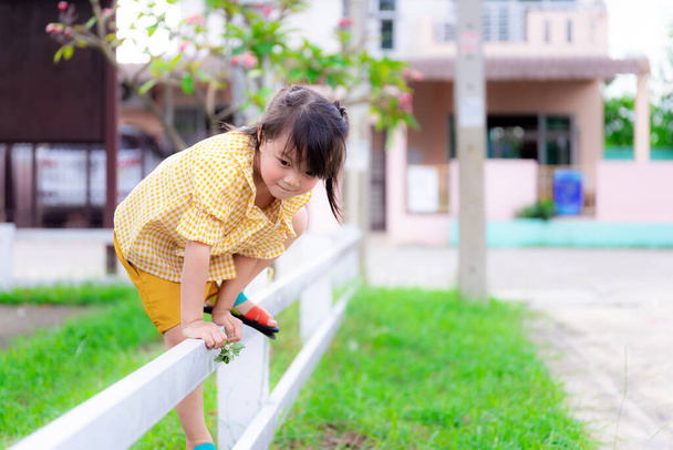 Active cute girl climbing white fence. In hand she holding flowers from the grass. Asian children wear yellow clothes. Child is fluent, smiling sweetly. Spring or summer. Asian girl 5 years old. - Photo, Image