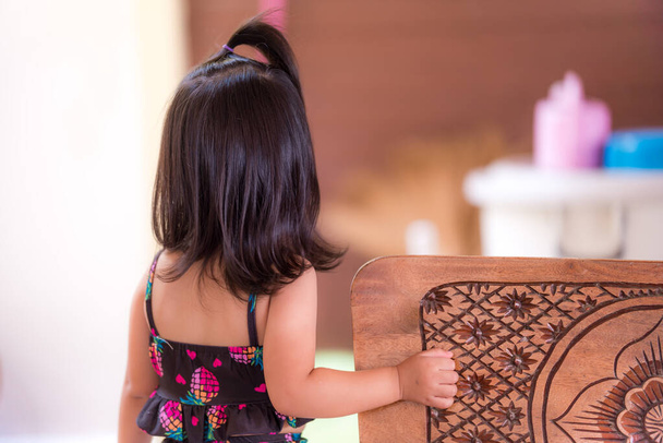 Le dos de la mignonne petite fille asiatique aux cheveux noirs à l'épaule. Un enfant en maillot de bain noir attendait sa mère devant la maison. Le gamin a 3 ans. - Photo, image