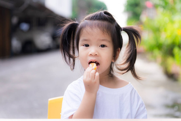 Schattig meisje bundelt haar en eet sinaasappels in haar mond. Kind droeg een wit shirt voor het huis. Achtergrond is een kleine openbare weg. Aziatische kinderen eten fruit, 3 jaar oud. - Foto, afbeelding