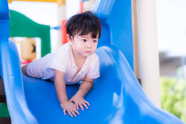 Asian toddler boy was face down to play on the blue slide. In hot day child sweats on his face. Child is worried about slipping downwards. Little boy face plants going down the slide head first. - Photo, Image