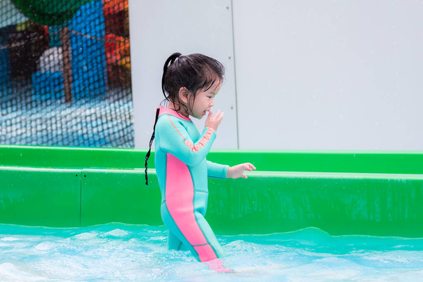 Sivunäkymä aasialaisesta lapsesta uimapuvussa mintunvihreässä. Lapsi pyyhki vettä kasvoilleen pelattuaan vesiliukumäkeä. Loma perheen kanssa vesipuistossa. Lasten leikkiä vedessä märkä, ikä 3 ja puoli vuotta vanha. - Valokuva, kuva