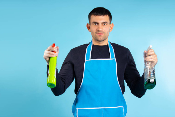 Serious man with cleaning sprays looking at the camera. Photo on a blue background with empty side space. The concept of gender stereotypes. - Zdjęcie, obraz