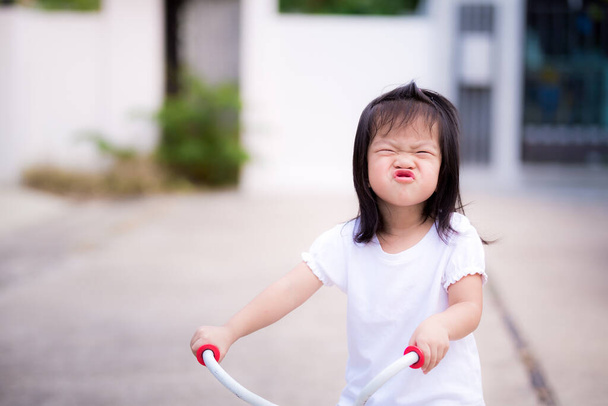 Preschool Asian little child girl make a playful face. Children wrapped up the mouth and  ride a bicycle. The kid wore a white dress in a good mood. Sweet smile in summer or spring. Baby 3 years old. - Photo, Image