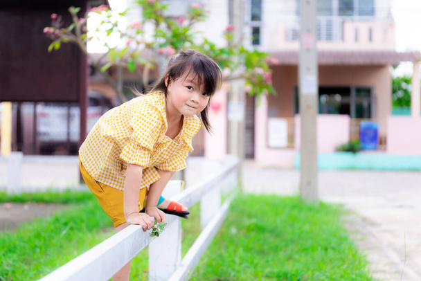 Pajkos lány átmászik a fehér kerítésen, hogy átkeljen a játszótérről, hogy hazamehessen. A fiatal lány izgatott és kedves mosollyal. Egy meleg nyáron vagy tavasszal. 5 éves gyermek. - Fotó, kép
