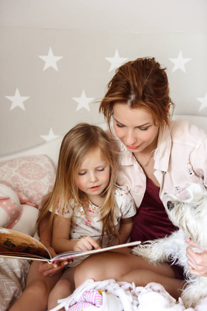 Mom reading a fairy tale in bed to a little girl. Near a dog west highland white terrier - Photo, image