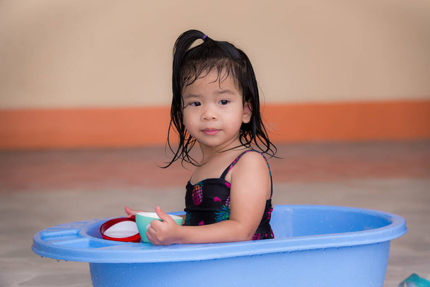 Tyttö uimapuvussa leikkimässä sinisellä altaalla talonsa edessä. Lapset pelaavat muovikuppeja, leluja ympäristöstä jokapäiväisessä elämässä. Söpö pieni lapsi, 3 vuotias. - Valokuva, kuva