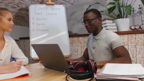 İngilizce sınıfı - bir kadın ve Afrika kökenli Amerikalı bir erkek - bir adam dizüstü bilgisayarın başında oturuyor ve gülüyor ve bir kadın notlar alıyor - Video, Çekim