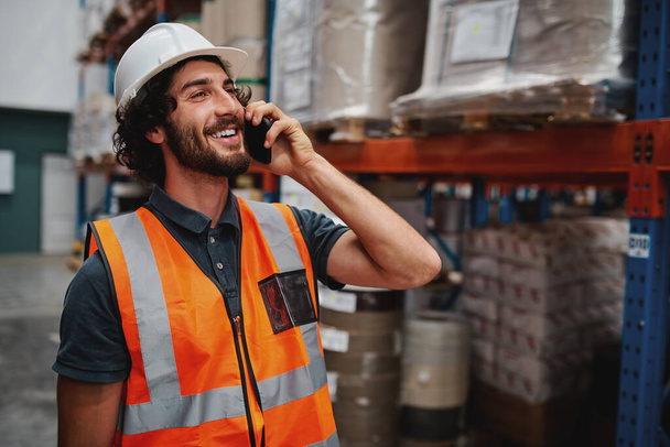 Happy bearded warehouse manager in conversation on mobile phone wearing white helmet and safety vest standing looking away - Foto, Imagem