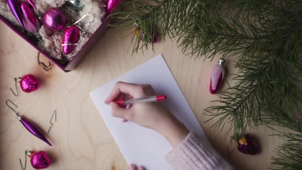 Vrouwelijke handen die een pen vasthouden en een brief schrijven aan de Kerstman. Doos met oude vintage kerstboom ornamenten en dennenbomen takken op lichte houten achtergrond. - Video