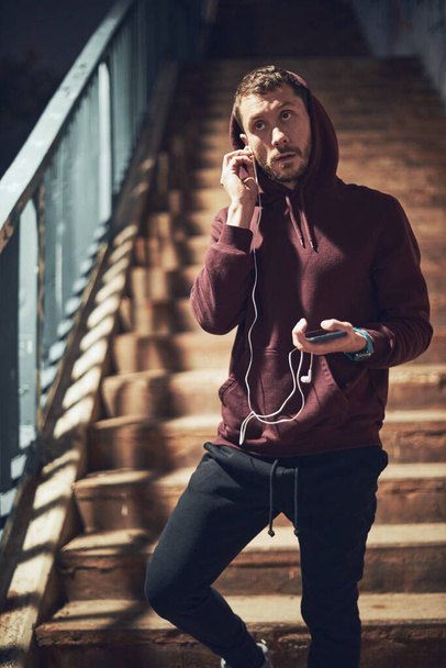 Athletic sporty man in hoodie sweatshirt making pause after training / exercising in urban area. - Фото, зображення