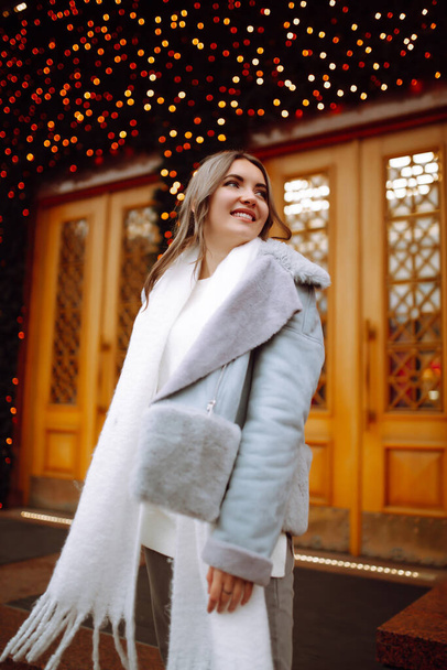Smiling woman in winter style clothes posing at festive street market.  Young woman enjoying winter moments. Festive Christmas fair, winter holidays concept. - Photo, image