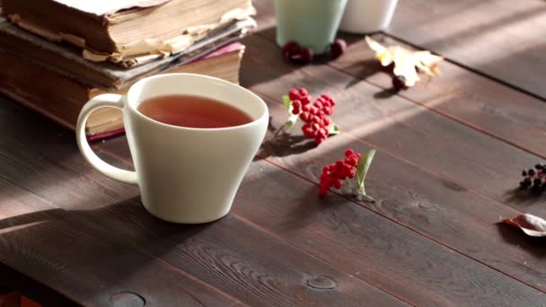 Fehér csésze tea gőzölög a fa asztalon. Régi könyvek, levelek, vörös bogyók. Őszi háttér - Felvétel, videó