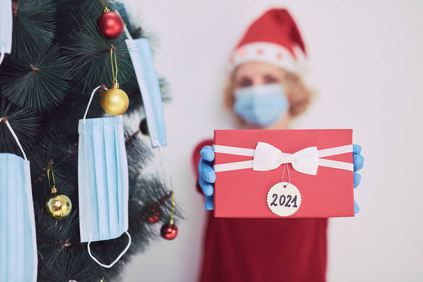 Vrouw met geschenkdoos aanwezig, kerstboom versieren met medische maskers tijdens viruspandemie 2020 / 2021. - Foto, afbeelding