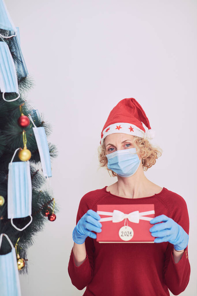Woman with gift box present, decorating christmas tree with medical masks during virus pandemic 2020 / 2021. - Fotografie, Obrázek