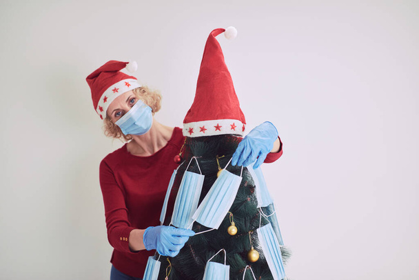 ウイルス流行中の医療マスクでクリスマスツリーを飾る女性2020/2021. - 写真・画像