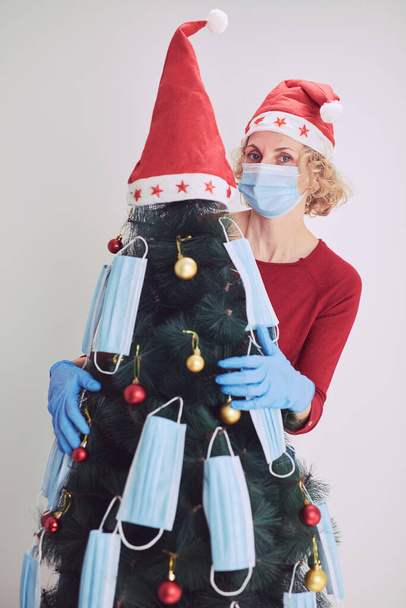 Γυναίκα διακόσμηση χριστουγεννιάτικο δέντρο με ιατρικές μάσκες κατά τη διάρκεια πανδημίας του ιού 2020 / 2021. - Φωτογραφία, εικόνα