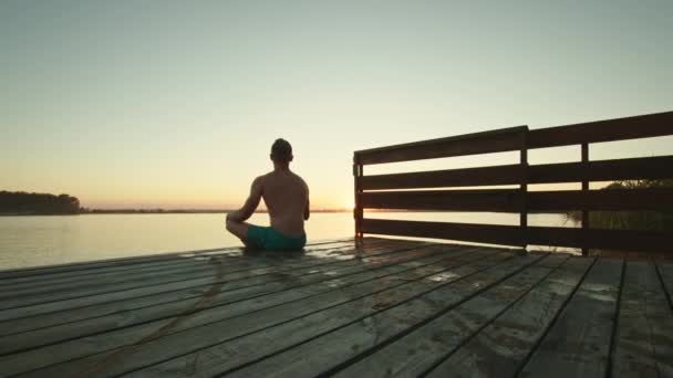 Um jovem está sentado à beira do cais e meditando. Belo pôr-do-sol. Descansa no lago. 4K - Filmagem, Vídeo