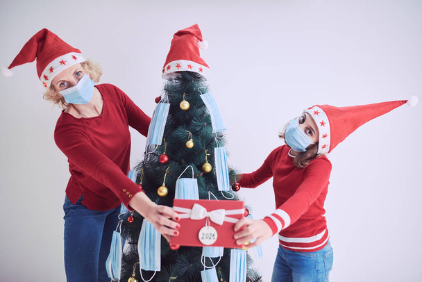 Moeder en dochter versieren kerstboom met medische maskers tijdens 2020 / 2021 virus pandemie. - Foto, afbeelding