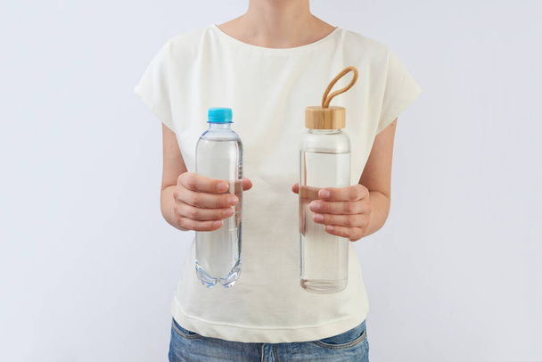 Two bottles of plastic and glass with clean natural water in a woman's hands against light grey background, copy space. Zero waste concept. Using a reusable glass bottle instead of single-use plastic. - Photo, image
