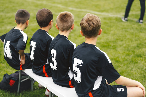 Group of young boys in sports football team sitting on white substitution bench and waiting for a chance to play game. Soccer match for junior footballers - Photo, Image