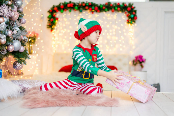 Щасливий хлопчик, одягнений як гном, сидить на підлозі у своїй вітальні під час Різдва. Дитина має різдвяний подарунок
. - Фото, зображення