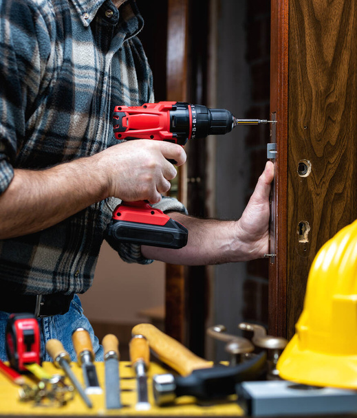 Close-up. Carpenter with an electric screwdriver fixes the lock of a wooden door. Construction industry.  - Photo, Image