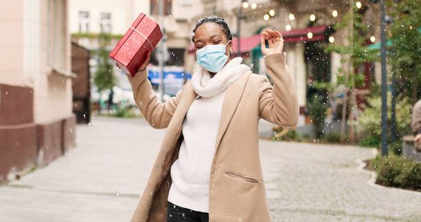 Happy beautiful smiling African American woman dancing in winter city with christmas gift in good mood. Portrait of joyful female in mask got holiday present on New years eve. Xmas concept - Photo, image