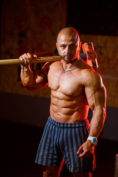 Silný atletický muž pózující se sportovním kladivem na pozadí tělocvičny. Silný kulturista s dokonalými svaly, rameny, bicepsy, tricepsy a hrudníkem. - Fotografie, Obrázek