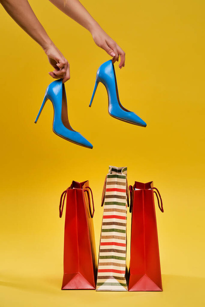Mani femminili che tengono le scarpe su uno sfondo giallo, borse della spesa sono in piedi nelle vicinanze. - Foto, immagini