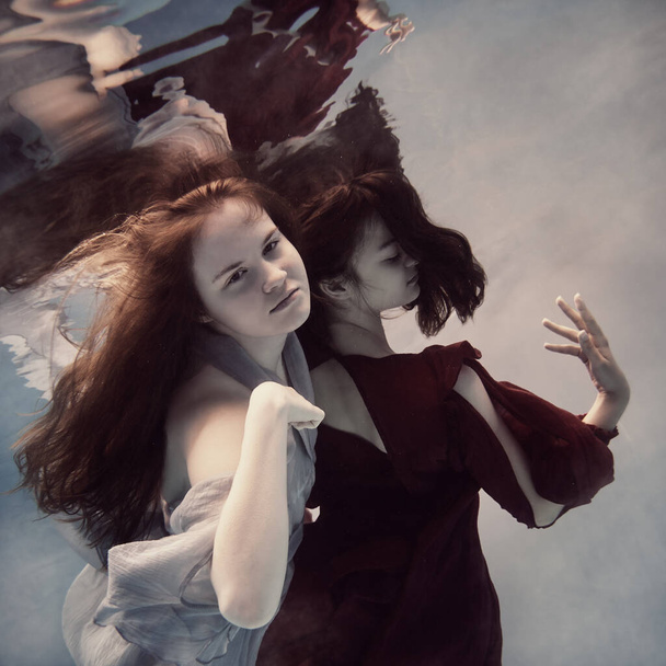 Two girls underwater in blue and red dresses with developing long hair seem to fly in weightlessness and interact with each other - Φωτογραφία, εικόνα