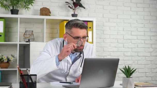 Lekarz bada pacjenta online wideo połączenie laptop kamera wygląda szkło powiększające - Materiał filmowy, wideo