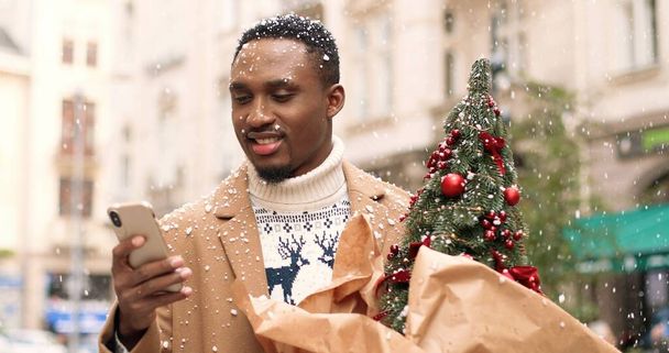 Close up portrait of young cheerful African American man standing in snowy xmas town and sending greeting on smartphone. Male with christmas small tree typing on cellphone. Holidays spirit concept - Foto, afbeelding