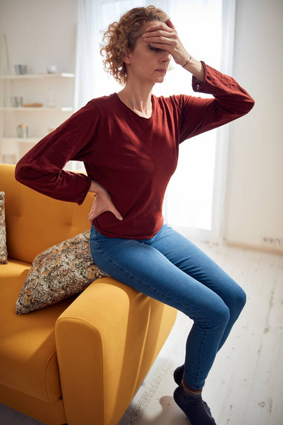 Femme avec douleur à la tête et à la hanche assise sur un canapé à la maison. - Photo, image