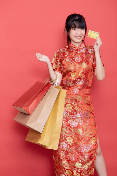 Portret Schattige jonge Aziatische vrouw dragen Chinese jurk met een boodschappentas en creditcard voor Chinees Nieuwjaar op een rode achtergrond - Foto, afbeelding