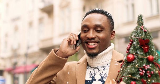 Handsome young happy African American man standing in winter city and chatting on smartphone and receiving holiday greetings. Male with small christmas tree talking on cellphone. Holiday concept - Foto, afbeelding