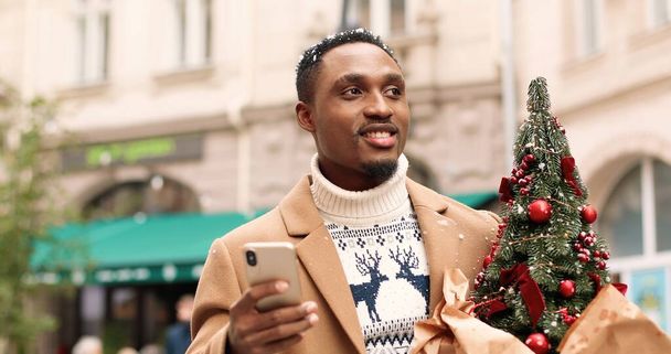 Handsome joyful African American man standing in city and texting on smartphone and receiving holiday greetings. Close up portrait of male with small christmas tree sends message on cellphone. - Fotó, kép