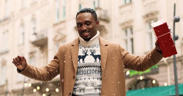 Happy handsome African American young man dancing in xmas city with christmas gift box in good mood. Portrait of joyful male got holiday present and making funny happy moves on street Holidays concept - Foto, Imagen