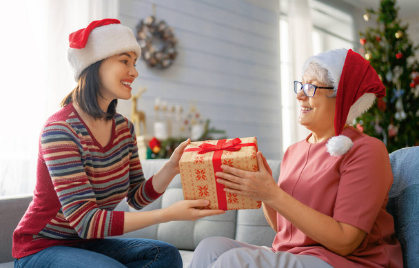 Merry Christmas and Happy Holidays! Senior mom and her adult daughter exchanging gifts. Having fun near tree indoors. Loving family with presents in room.  - Φωτογραφία, εικόνα