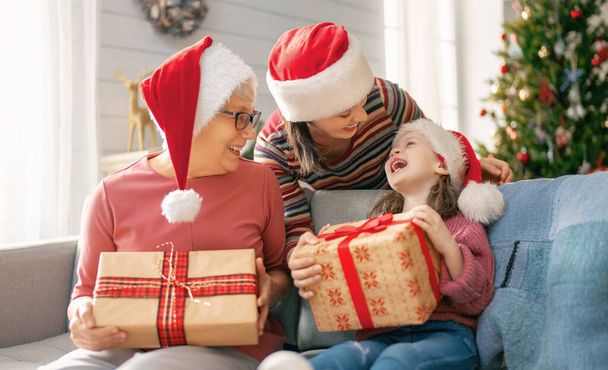 Merry Christmas and Happy Holidays! Cheerful kid presenting gifts to mom and granny. Parents and little child having fun near tree indoors. Loving family with presents in room. - Photo, Image
