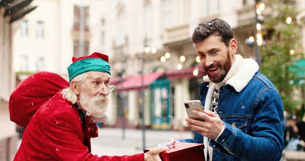 Close up portrait of happy Caucasian male standing in city and typing on smartphone while meeting old man dressed like Santa Claus who is giving him xmas present. Guy taking selfie photo on cellphone - Photo, Image