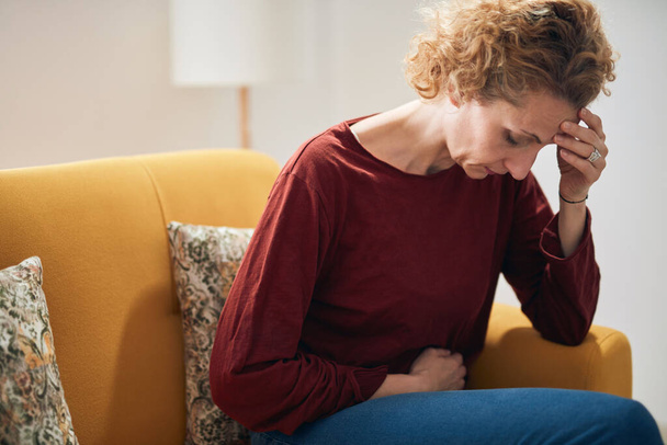 Frau mit Bauchschmerzen und Kopfschmerzen sitzt zu Hause auf einer Couch. - Foto, Bild