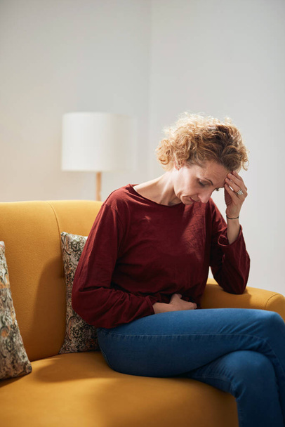 Donna con mal di stomaco e mal di testa problema seduto su un divano a casa. - Foto, immagini