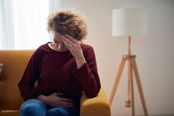 Femme avec des douleurs à l'estomac et des maux de tête problème assis sur un canapé à la maison. - Photo, image